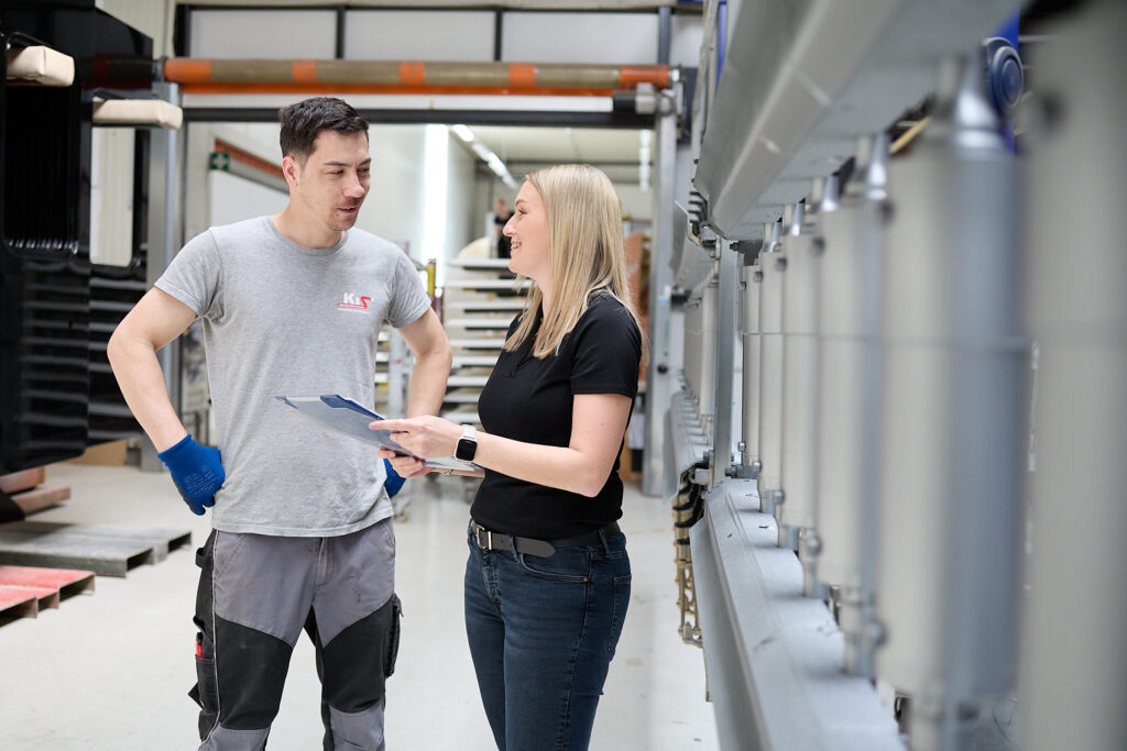 Zwei Personen stehen in einem industriellen Arbeitsraum und diskutieren über ein Dokument. Der Mann trägt Arbeitshandschuhe und die Frau hält ein Klemmbrett in der Hand. Im Hintergrund sind Maschinen und Geräte zu sehen. LAC Berger Industrielackierung