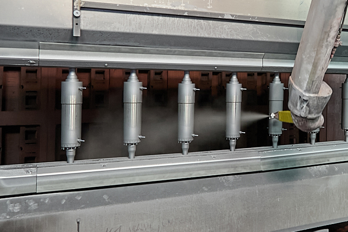 In einer industriellen Produktionsanlage werden mehrere zylindrische Metallteile aufgereiht und von einer automatischen Spritzdüse beschichtet. LAC Berger Industrielackierung